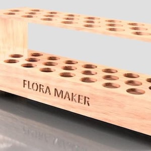 Op de afbeelding: Een houten standaard met 30 ronde gaten om buizen of flesjes in te bewaren. De standaard heeft twee planken en is gelabeld met "FLORA MAKER".