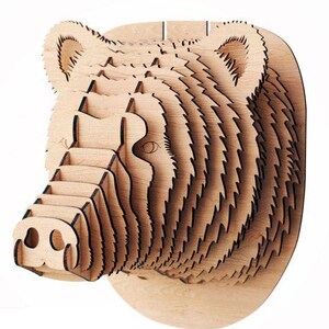Può includere: Una scultura di testa di orso in legno con un design a strati. La scultura è realizzata con più pezzi di legno impilati uno sopra l'altro per creare un effetto 3D. La testa dell'orso è rivolta a sinistra e i suoi occhi sono visibili. La scultura è di colore marrone chiaro.