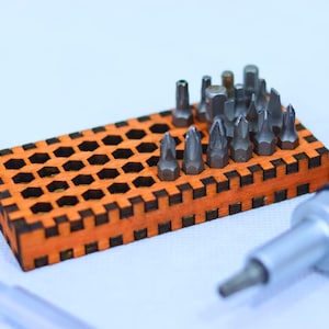 Op de afbeelding: Een set schroevendraaierbits in een houten houder met een zeshoekig rasterpatroon. De bits zijn in rijen gerangschikt en zijn gemaakt van metaal. Een schroevendraaierhandvat is ook zichtbaar op de afbeelding.
