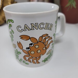 May include: White ceramic mug with a brown crab illustration and the word "Cancer" in yellow lettering. The mug has green leafy designs around the crab.
