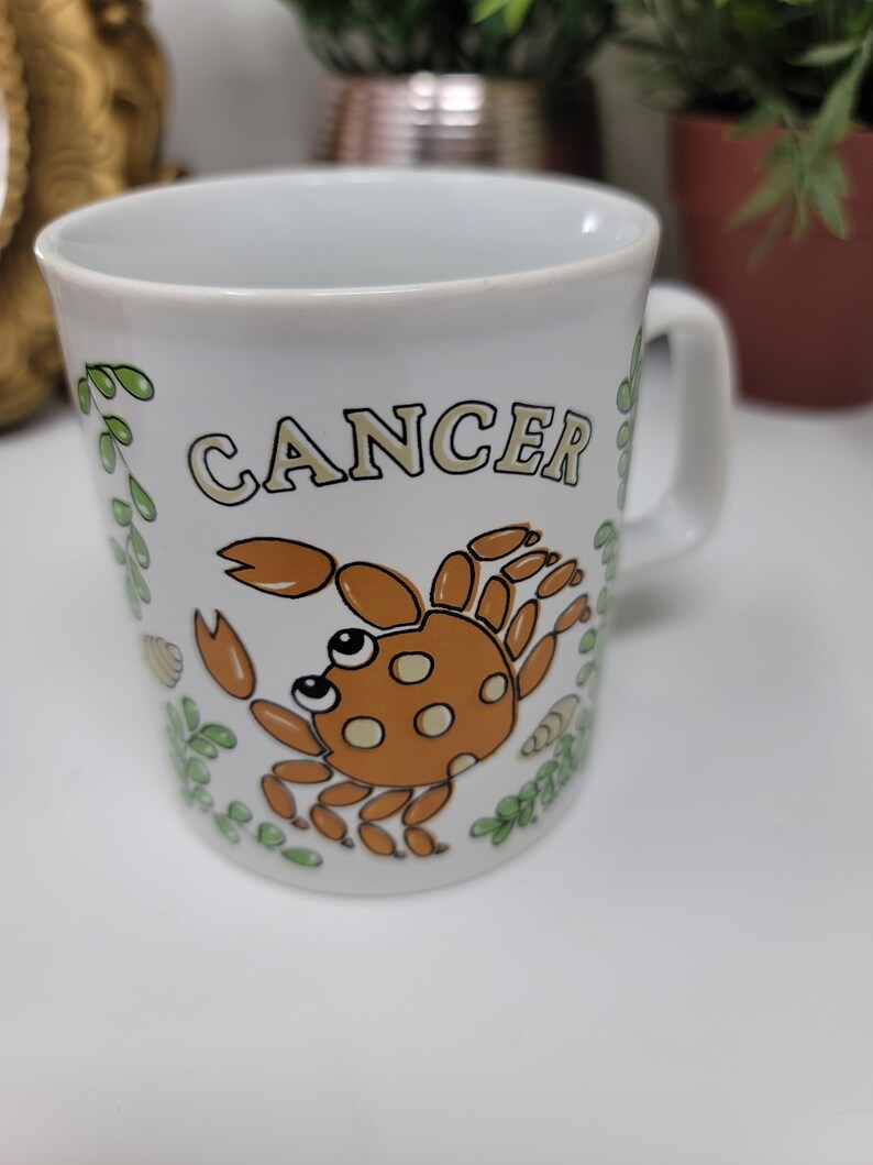 May include: White ceramic mug with a brown crab illustration and the word "Cancer" in yellow lettering. The mug has green leafy designs around the crab.