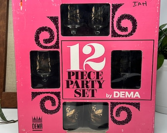 May include: A pink box with a black and gold 12 piece party set of glasses by Dema. The box has a crown logo and the text "12 Piece Party Set by Dema".