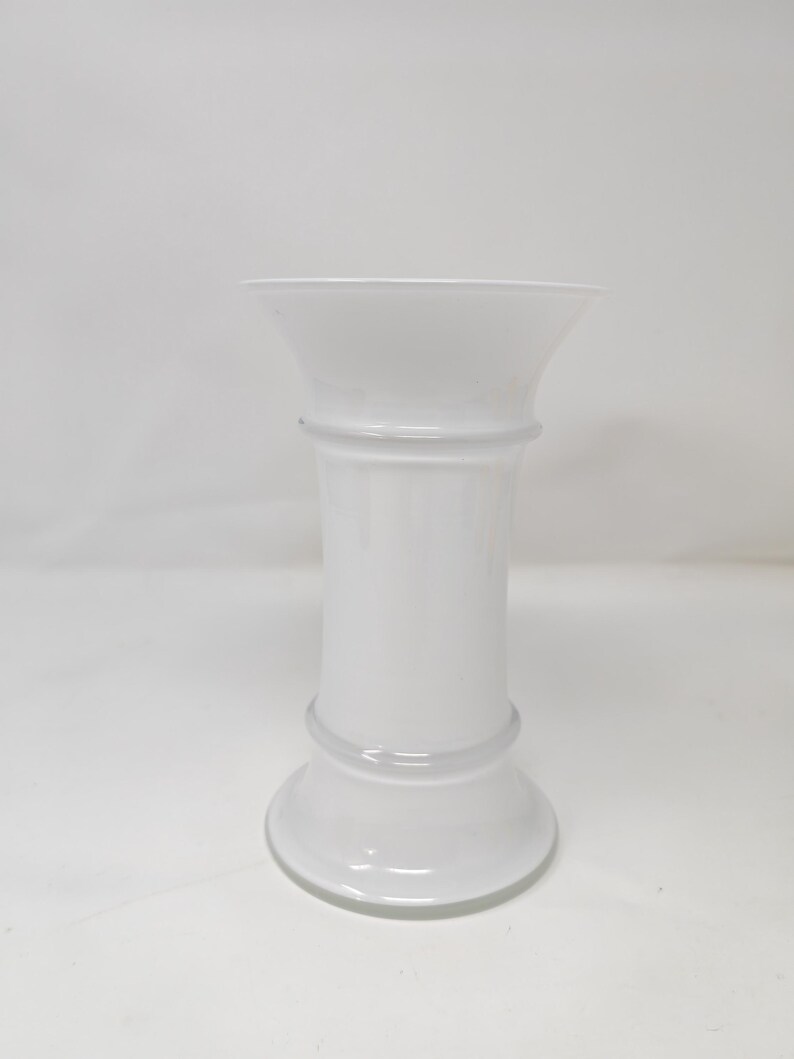 May include: A white, opaque glass vase with a flared top and a rounded base. The vase has a cylindrical body with two horizontal bands. The overall design is simple and elegant.