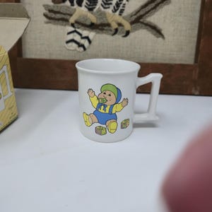 Pode incluir: Caneca de cerâmica branca com um bebê de desenho animado em macacão azul, chapéu verde e sapatos amarelos. O bebê é cercado por blocos ABC. A caneca tem uma alça quadrada, provavelmente vintage.