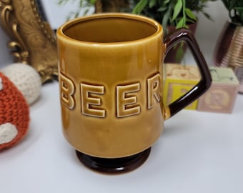 May include: A brown ceramic mug with a brown handle. The mug has the word "BEER" embossed on the front.