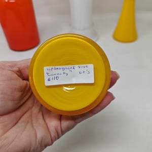 May include: A close-up of a yellow glass vase base with a handwritten label. The label reads "Holmegaard Vase Carnaby 60's". Other glass vases in orange, white, and yellow are in the background.