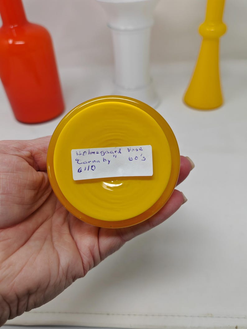 May include: A close-up of a yellow glass vase base with a handwritten label. The label reads "Holmegaard Vase Carnaby 60's". Other glass vases in orange, white, and yellow are in the background.