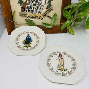 May include: Two white ceramic plates with gold trim and a raised design. One plate features a girl in a yellow dress and a red hat, the other features a boy in a blue outfit. Both plates have a gold floral design around the edge.