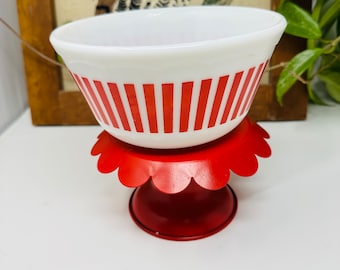 May include: A white milk glass bowl with red stripes sits on a red metal cake stand with scalloped edges.