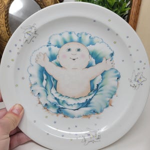 May include: White ceramic plate with a baby illustration at the centre, framed by blue and white floral details. The rim features small star and bunny illustrations. The plate is held by a hand.