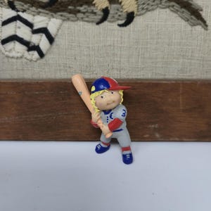 Könnte beinhalten: Kleine Figur eines Baseballspielers in grauer Uniform mit roten und blauen Akzenten, der einen Holzschläger hält. Der Spieler trägt eine blau-rote Kappe und blaue Schuhe. Der Hintergrund zeigt eine gestickte Eule.