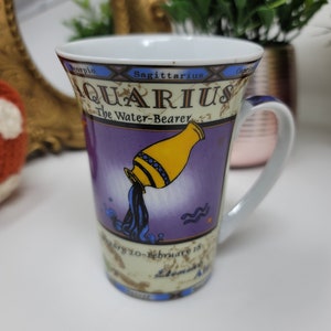 Op de afbeelding: Witte keramische Aquarius-mok met een paarse achtergrond en een gele kruik die blauw water giet. De mok toont de tekst "Aquarius The Water-Bearer" en de data "20 januari-18 februari".