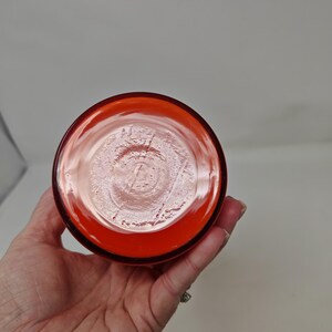 May include: A bottom view of a clear, orange-colored glass. The glass has a circular shape with a thick rim. The base of the glass has a textured pattern. The glass is held by a hand.