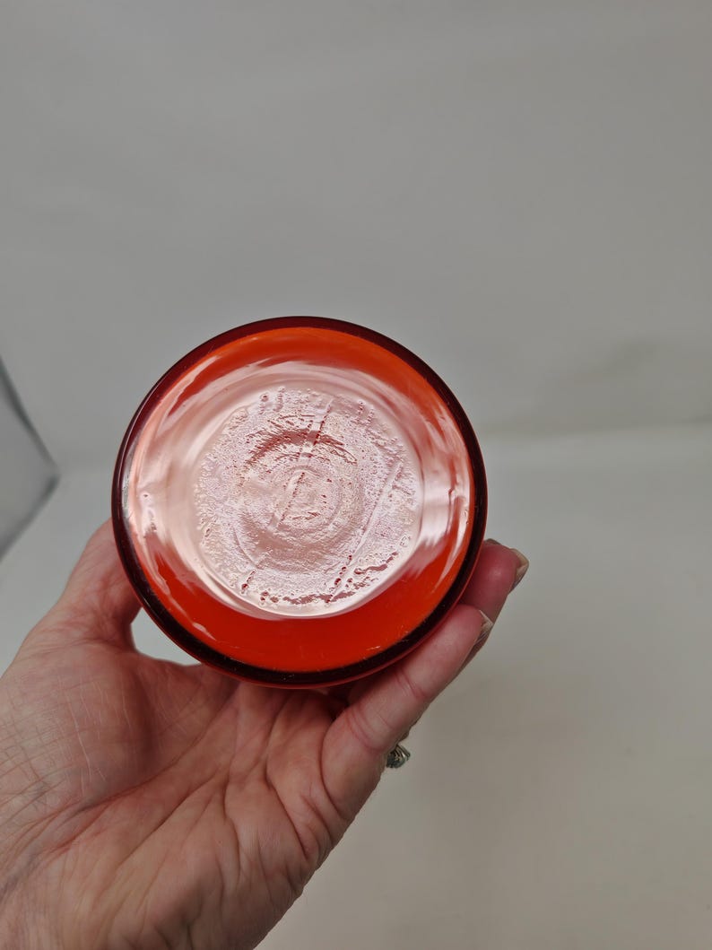 May include: A bottom view of a clear, orange-colored glass. The glass has a circular shape with a thick rim. The base of the glass has a textured pattern. The glass is held by a hand.