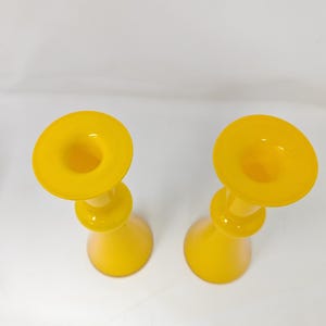 May include: Two yellow glass candle holders with a rounded base and a flared top. The candle holders are made of a translucent glass material and are set against a white background. The design is simple and elegant.