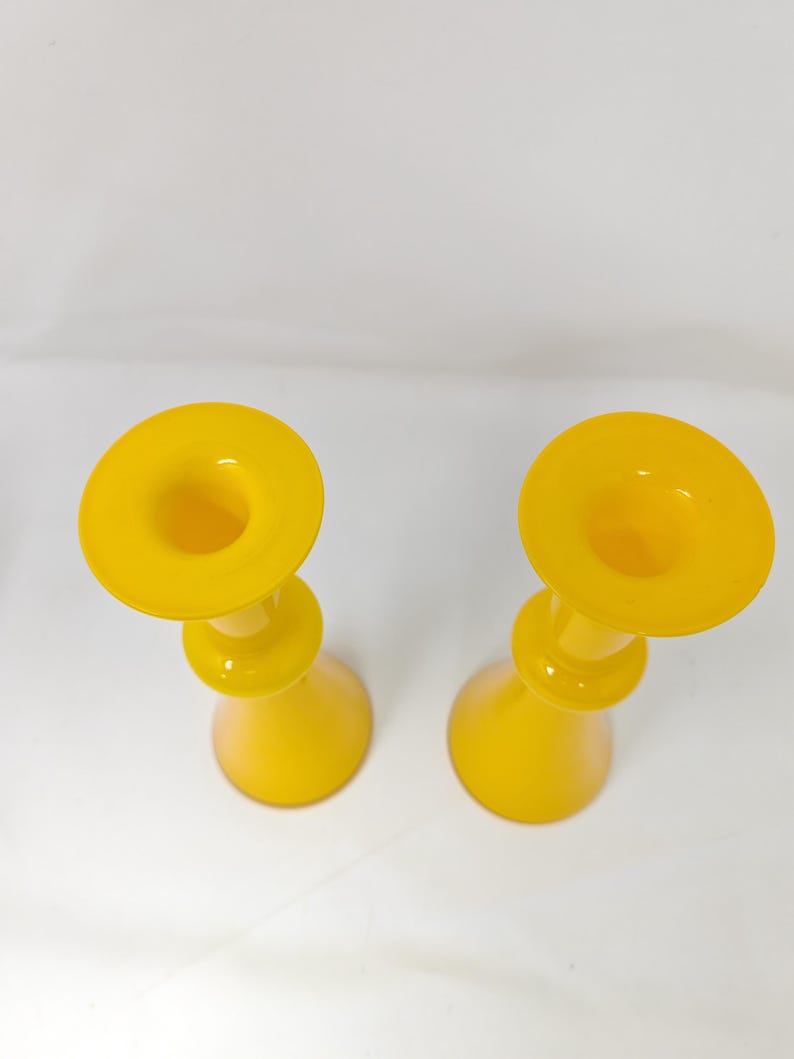 May include: Two yellow glass candle holders with a rounded base and a flared top. The candle holders are made of a translucent glass material and are set against a white background. The design is simple and elegant.