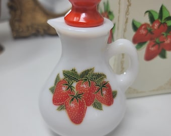 Puede incluir: Una crema de cerámica blanca con un tapón rojo. La crema tiene un asa y un diseño de fresas. El tapón es una esfera roja brillante. La crema es probablemente vintage y parte de un conjunto.