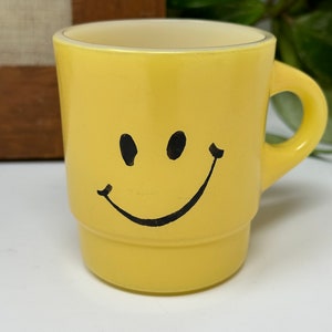 Puede incluir: Una taza de cerámica amarilla con una carita sonriente negra pintada en la parte delantera.