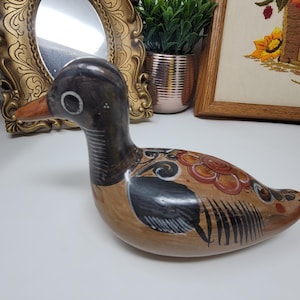 May include: A hand-painted ceramic duck figurine. The duck has a brown body with black and red floral designs, a black head, and an orange beak. The figurine is on a white surface.