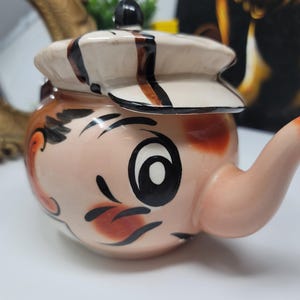 May include: A vintage ceramic teapot with a whimsical face design. The teapot has a light pink base with painted details in black, brown, and red. The lid is shaped like a cap with brown and black stripes. The spout and handle are light pink.