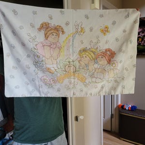 Pode incluir: Fronha branca com uma ilustração colorida de Cabbage Patch Kids. O design inclui três bonecas, um arco-íris, borboletas e detalhes florais. As palavras "Cabbage Patch Kids" também são visíveis.