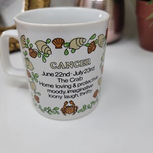 May include: White ceramic mug with a green, brown, and orange design featuring a crab and seashells. The mug has the text "CANCER June 22nd - July 23rd The Crab Home loving & protective moody, imaginative loony laugh, thrifty."