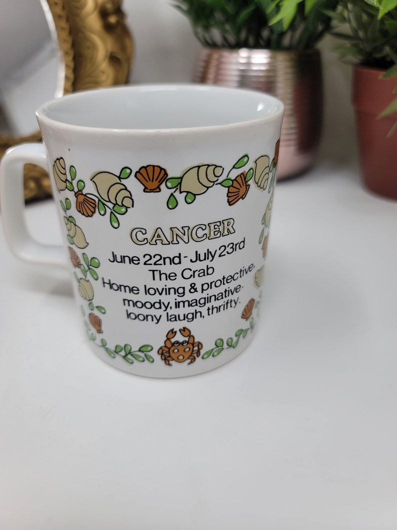 May include: White ceramic mug with a green, brown, and orange design featuring a crab and seashells. The mug has the text "CANCER June 22nd - July 23rd The Crab Home loving & protective moody, imaginative loony laugh, thrifty."