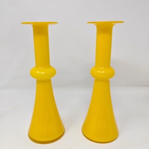 May include: Two bright yellow glass vases. Each vase has a flared base, a bulbous midsection, and a tall, cylindrical neck that flares out at the top. The vases are identical in shape and color, and stand against a white background.