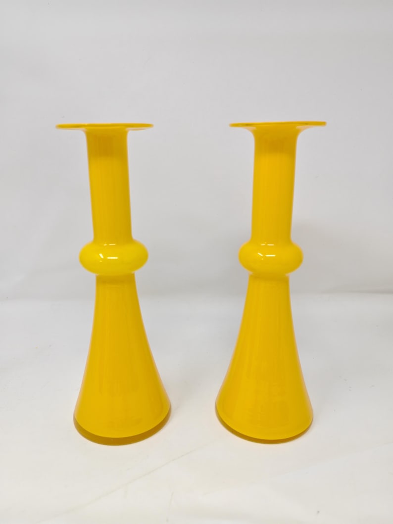 May include: Two bright yellow glass vases. Each vase has a flared base, a bulbous midsection, and a tall, cylindrical neck that flares out at the top. The vases are identical in shape and color, and stand against a white background.