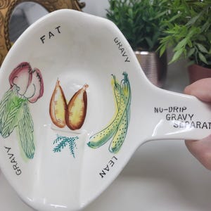 Puede incluir: Salsera de cerámica blanca con una sección dividida para grasa, magra y salsa. La salsera está decorada con una verdura verde de hoja, una flor roja y blanca, dos patatas marrones y tres judías verdes. Las palabras "FAT", "LEAN", "GRAVY" y "NU-DRIP GRAVY SEPARAT" están impresas en la salsera.