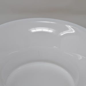May include: A white ceramic bowl with a wide, shallow design. The bowl has a smooth, glossy finish and a slightly curved rim. The interior of the bowl is also white and reflects light. The bowl is likely for serving or display.