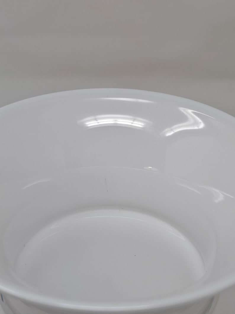 May include: A white ceramic bowl with a wide, shallow design. The bowl has a smooth, glossy finish and a slightly curved rim. The interior of the bowl is also white and reflects light. The bowl is likely for serving or display.