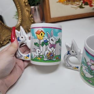Może przedstawiać: Białe ceramiczne kubki wielkanocne z uchwytami w kształcie królików i kolorowymi ilustracjami. Kubki przedstawiają scenę z królikami z budką dla ptaków, kwiatami i królikiem trzymającym koszyk. Uchwyty mają kształt królików z niebieskimi kokardkami.