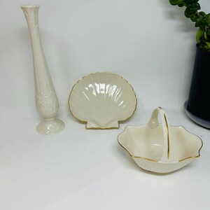 May include: A set of cream-coloured ceramic ornaments. It features a tall vase, a shell-shaped dish, and a handled basket, all with gold edging. The vase has a floral pattern.