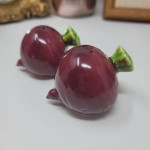 May include: A pair of ceramic salt and pepper shakers, each shaped like a red beetroot with a green leafy top. The shakers have a glossy finish and small holes. A delightful addition to any kitchen or dining table setting.