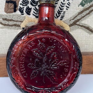 Puede incluir: Una botella redonda de vidrio rojo rubí con un tapón plateado. La botella presenta texto en relieve y un diseño de hoja de acebo. El texto dice "CHRISTMAS 1976". Es probable que sea un objeto de colección navideño vintage.