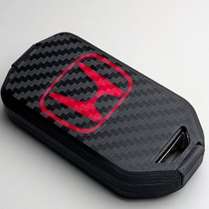 May include: Black key fob cover with a carbon fiber texture and a red Honda logo. The fob has a rectangular shape with rounded edges and a key ring attachment.