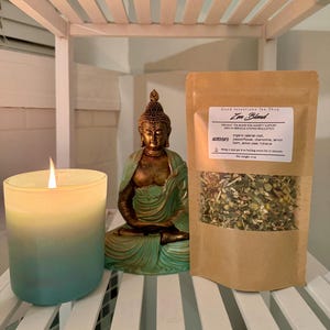 May include: A lit candle in a blue and yellow gradient glass jar, a seated Buddha statue, and a package of Zen Blend tea are arranged on a white shelf. The tea package has a label with the text "Good Intentions Tea Shop" and "Zen Blend".