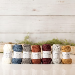 May include: A collection of seven yarn skeins in various colors, including cream, blue, gray, rust, burgundy, silver, and gold. Each skein has a label that reads "Feels Like Alpaca". The yarns are arranged on a white surface against a white wooden backdrop.