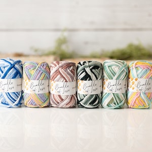 May include: Six colorful yarn bundles are displayed, each wrapped with a label that reads "Bundle of Love." The yarn colors include shades of blue, purple, pink, green, and yellow. The background is a blurred white surface with greenery.