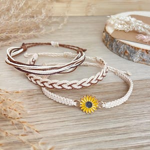 May include: A set of three bracelets featuring a sunflower charm with yellow petals and a dark centre, a braided cord bracelet in brown and cream, and a multi-strand cord bracelet in brown and cream, all crafted from woven string materials.
