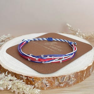 May include: A multi-strand bracelet featuring thin red, white, and blue cord strings accented with small, clear, transparent beads. The bracelet includes an adjustable sliding knot closure and is displayed on a brown rectangular card.