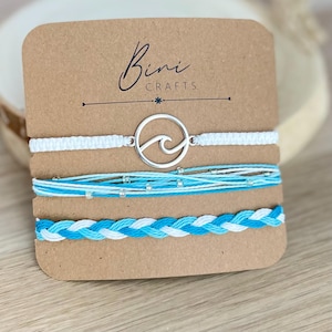 May include: A set of three bracelets on a brown card labelled Bini Crafts. One bracelet features a silver wave charm on a white cord, another consists of multiple light blue and white strands with small beads, and the third is a braided cord.