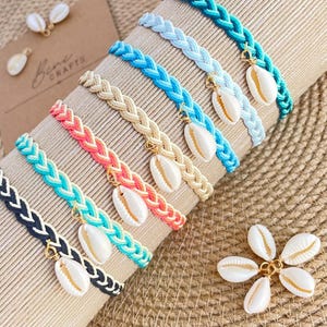 May include: A collection of braided cord bracelets in various colors including teal, light blue, beige, pink, and black. Each bracelet features a single white cowrie shell charm attached with a gold-tone metal jump ring. A cluster of five loose cowrie shells is also visible.