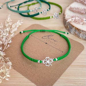 Puede incluir: Una pulsera de cordón trenzado de color verde brillante con un dije de trébol de cuatro hojas plateado y pequeñas cuentas plateadas. La pulsera se presenta sobre una tarjeta marrón con el texto Bini CRAFTS impreso en tinta negra.