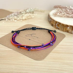 May include: A multi-strand bracelet featuring alternating black, orange, and purple threads accented with small, faceted purple beads. The bracelet is displayed on a square brown card with rounded corners that features the text Bini CRAFTS.