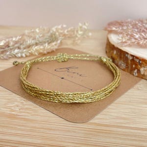 Könnte beinhalten: Ein goldfarbenes Kordelarmband aus mehreren dünnen, metallischen Strängen, die miteinander verflochten sind. Das Armband liegt auf einer braunen rechteckigen Karte mit dem in Schwarz gedruckten Text Bini CRAFTS, einer Linie und einem Stern.
