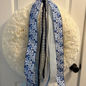 May include: A white wreath with blue and white patterned ribbon, blue lace, and white ribbon hanging from it.