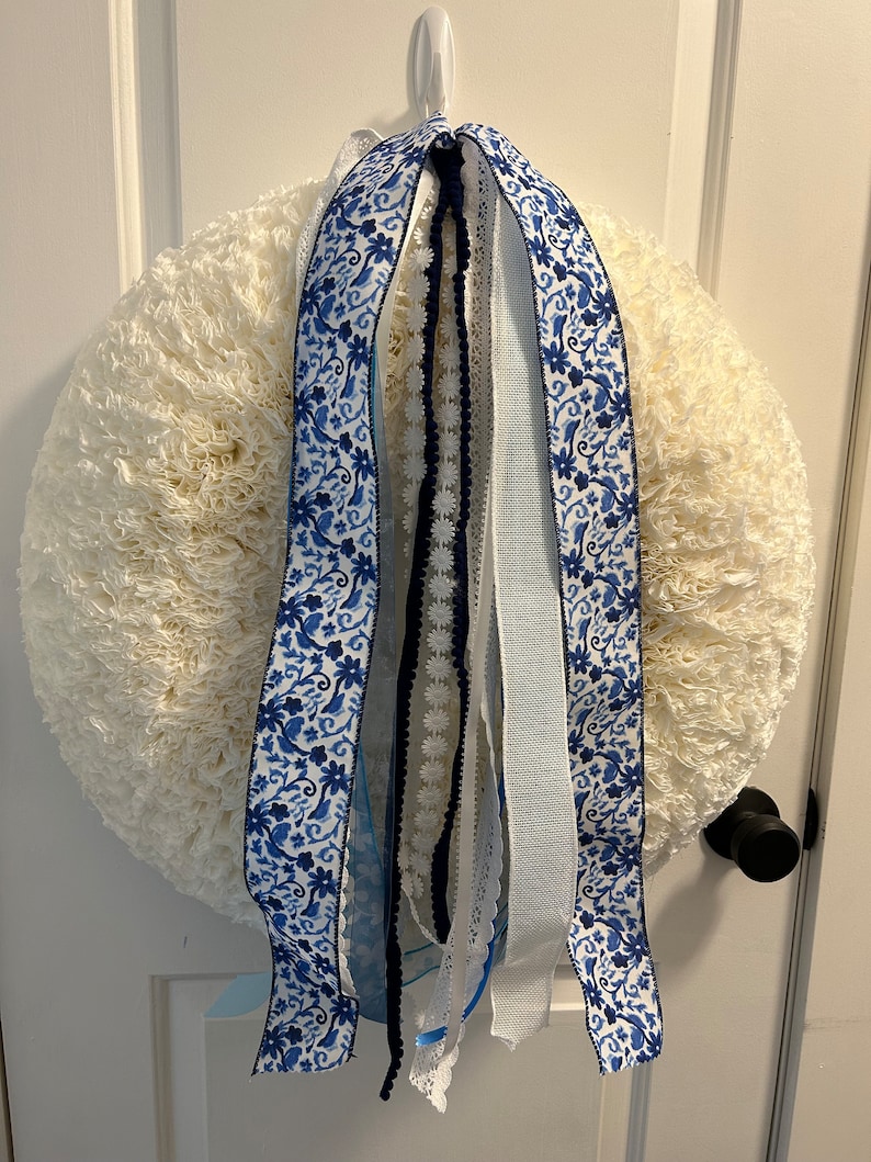 May include: A white wreath with blue and white patterned ribbon, blue lace, and white ribbon hanging from it.