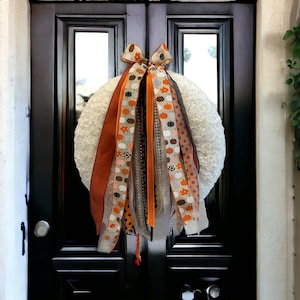 May include: A white wreath with orange, black, and white ribbon streamers with a pumpkin pattern. The wreath is hanging on a black door.